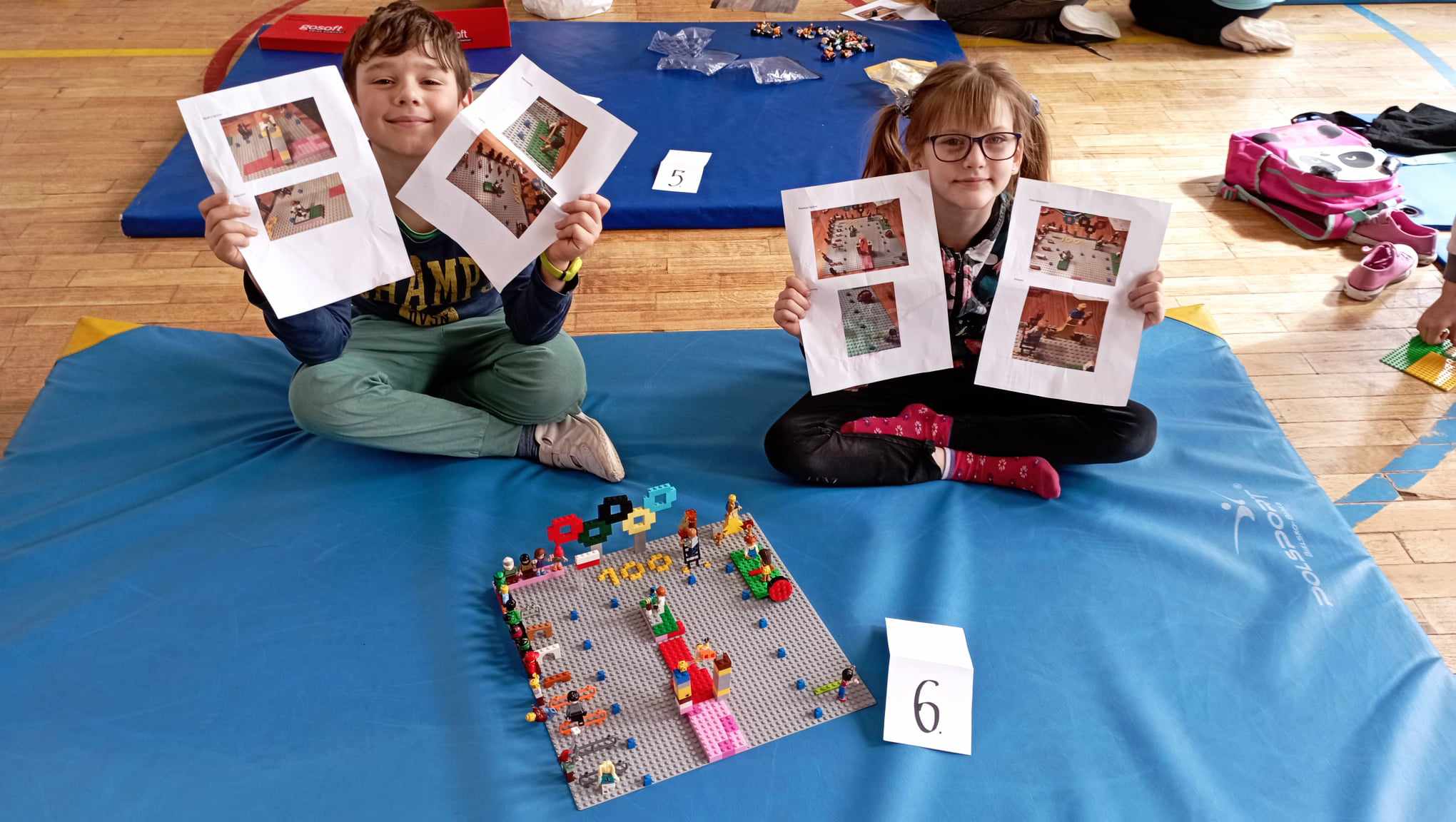 W sali gimnastycznej na niebieskim materacu siedzi dwoje uczniów z klasy II. Przed nimi stoi ich budowla z klocków LEGO i etykietka z cyfrą 6. Uczniowie trzymają w rękach kartki ze swoim projektem budowli.