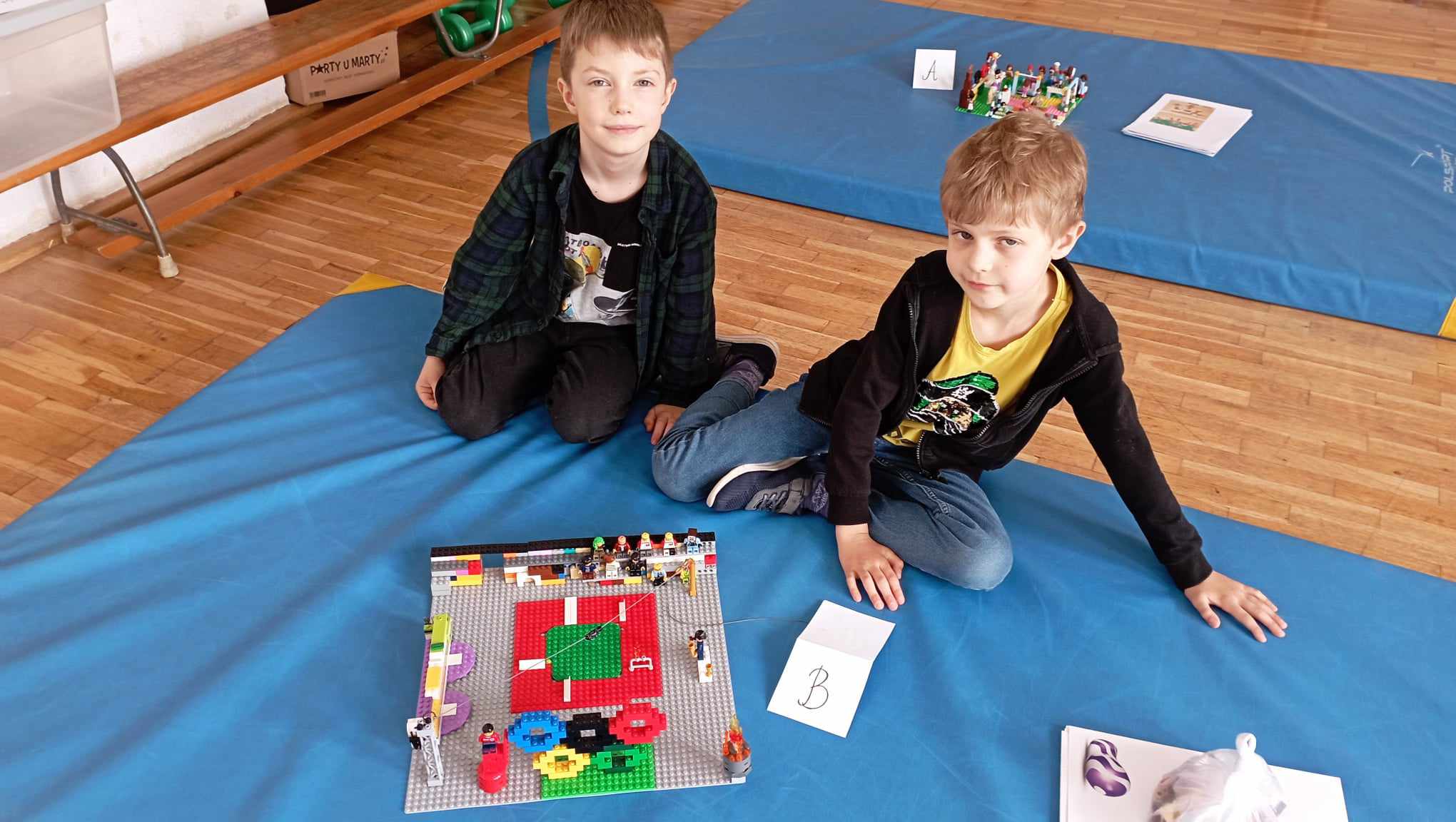 W sali gimnastycznej na niebieskim materacu siedzi dwoje uczniów z klasy 0. Przed nimi stoi ich budowla z klocków LEGO i etykietka z literą B, w tle inny materac z budowlą z klocków LEGO.