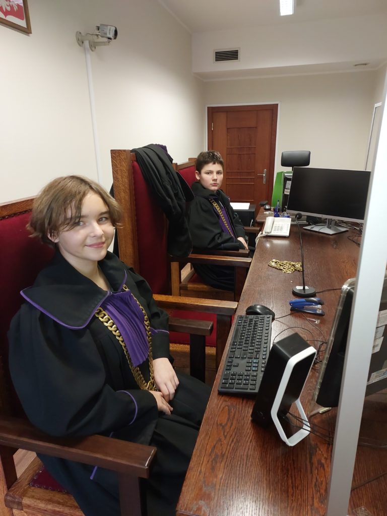 W sali rozpraw na krzesłach przy biurku siedzą: dziewczynka i chłopiec przebrani za sędziów. Na biurku znajduje się komputer i telefon.