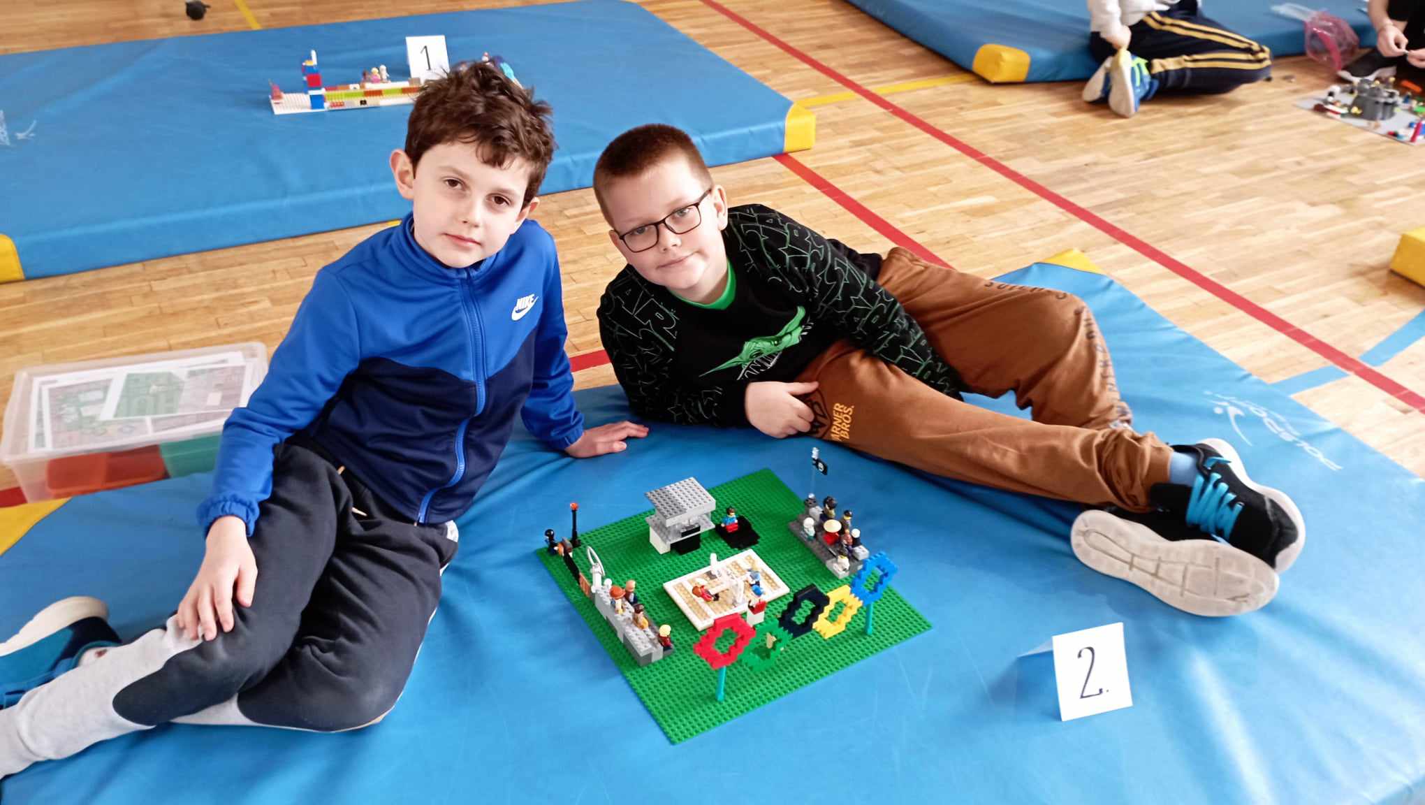 W sali gimnastycznej na niebieskim materacu siedzi dwoje uczniów z klasy II. Przed nimi stoi ich budowla z klocków LEGO i etykietka z cyfrą 2. W tle dwa inne materace z klockami