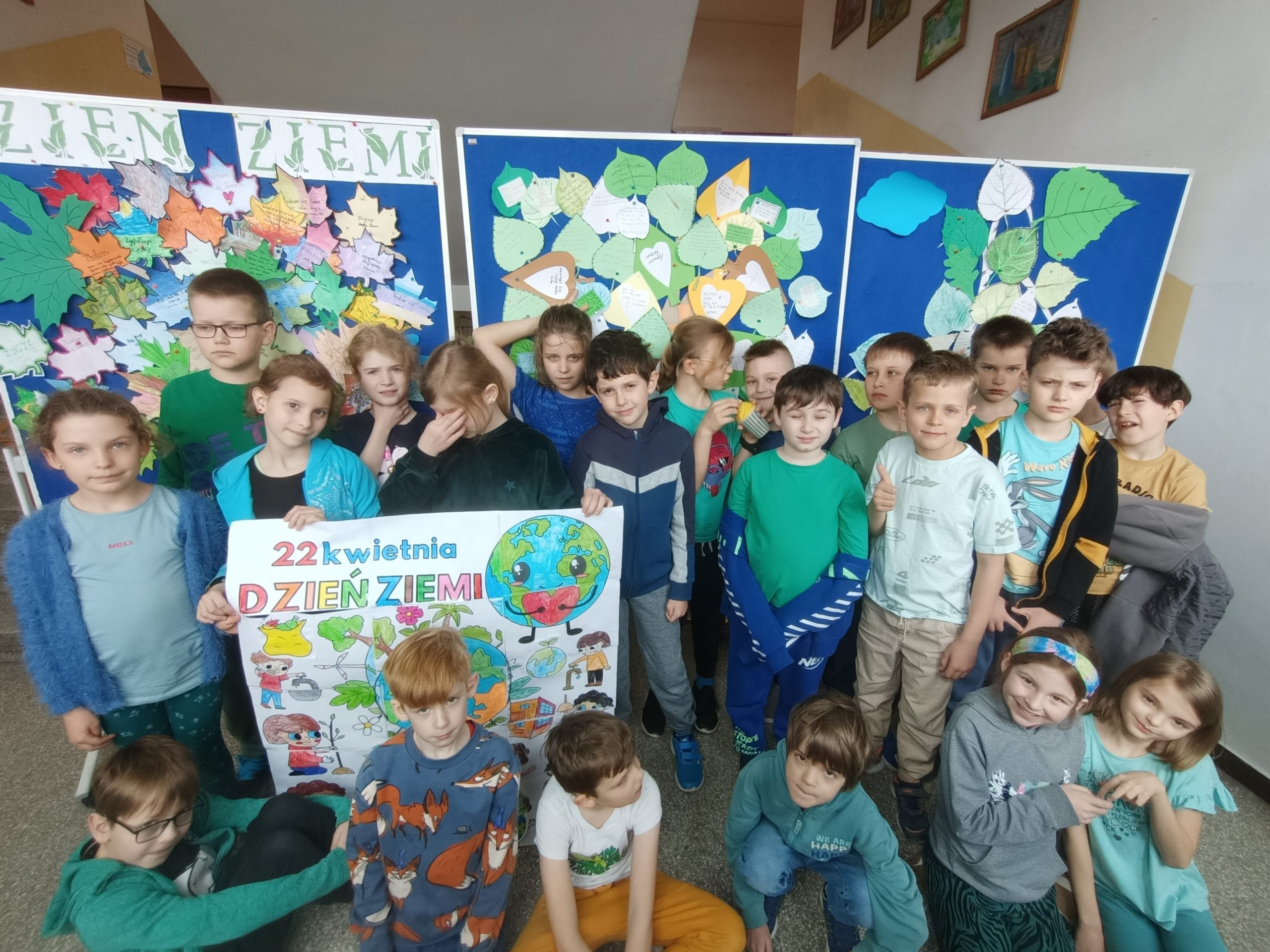 Grupa dzieci ustawiona w dwóch rzędach na szkolnym korytarzu. Dzieci w pierwszym rzędzie klęczą na podłodze, w drugim stoją na tle trzech tablic przedstawiających zrobione z papieru drzewa z liśćmi i napis "Dzień Ziemi". Troje dzieci trzyma plakat o tematyce ekologicznej.