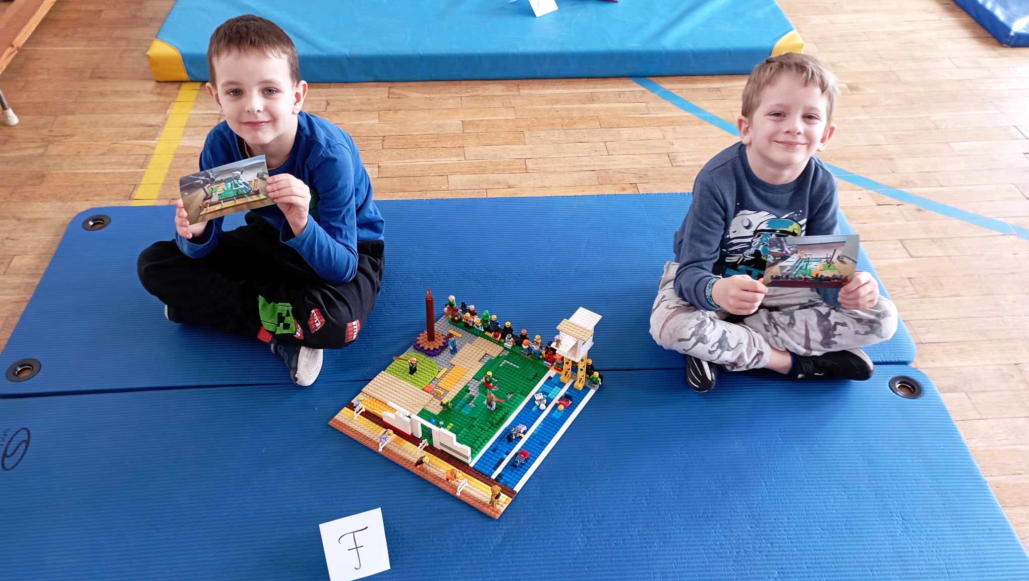 W sali gimnastycznej na niebieskim materacu siedzi dwoje uczniów z klasy 0, w rękach trzymają zdjęcia z projektem swojej budowli. Przed nimi stoi ich budowla z klocków LEGO i etykietka z literą F.