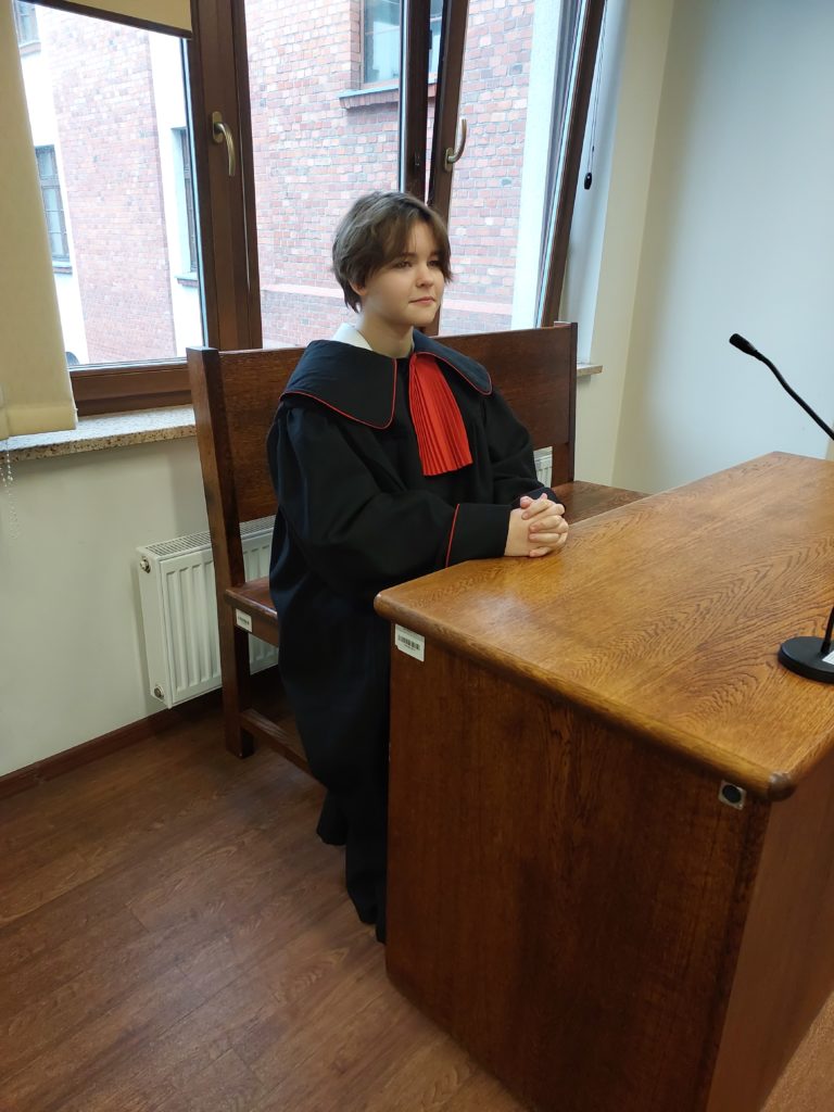 W Sali rozpraw przy biurku siedzi dziewczynka przebrana za prokuratora.