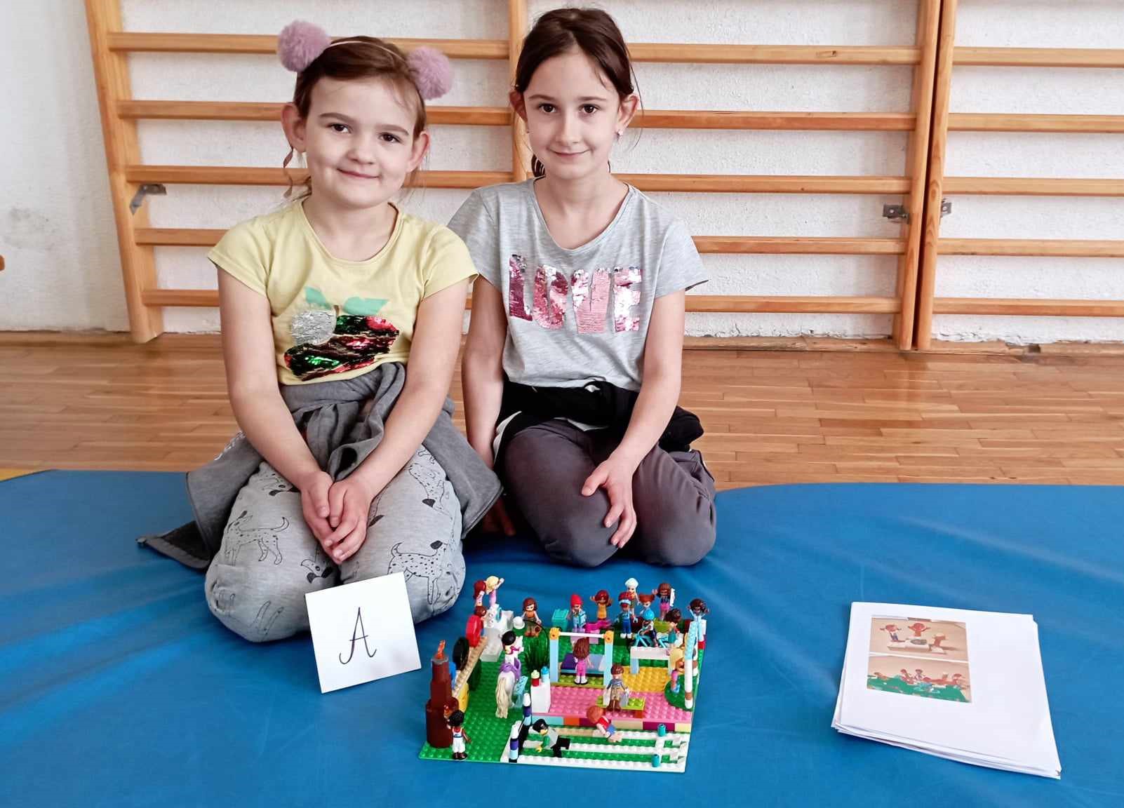 W sali gimnastycznej na niebieskim materacu siedzą dwie dziewczynki z klasy 0. Przed nimi stoi ich budowla z klocków LEGO i etykietka z literą A, obok kartki z projektem budowli. W tle drabinki