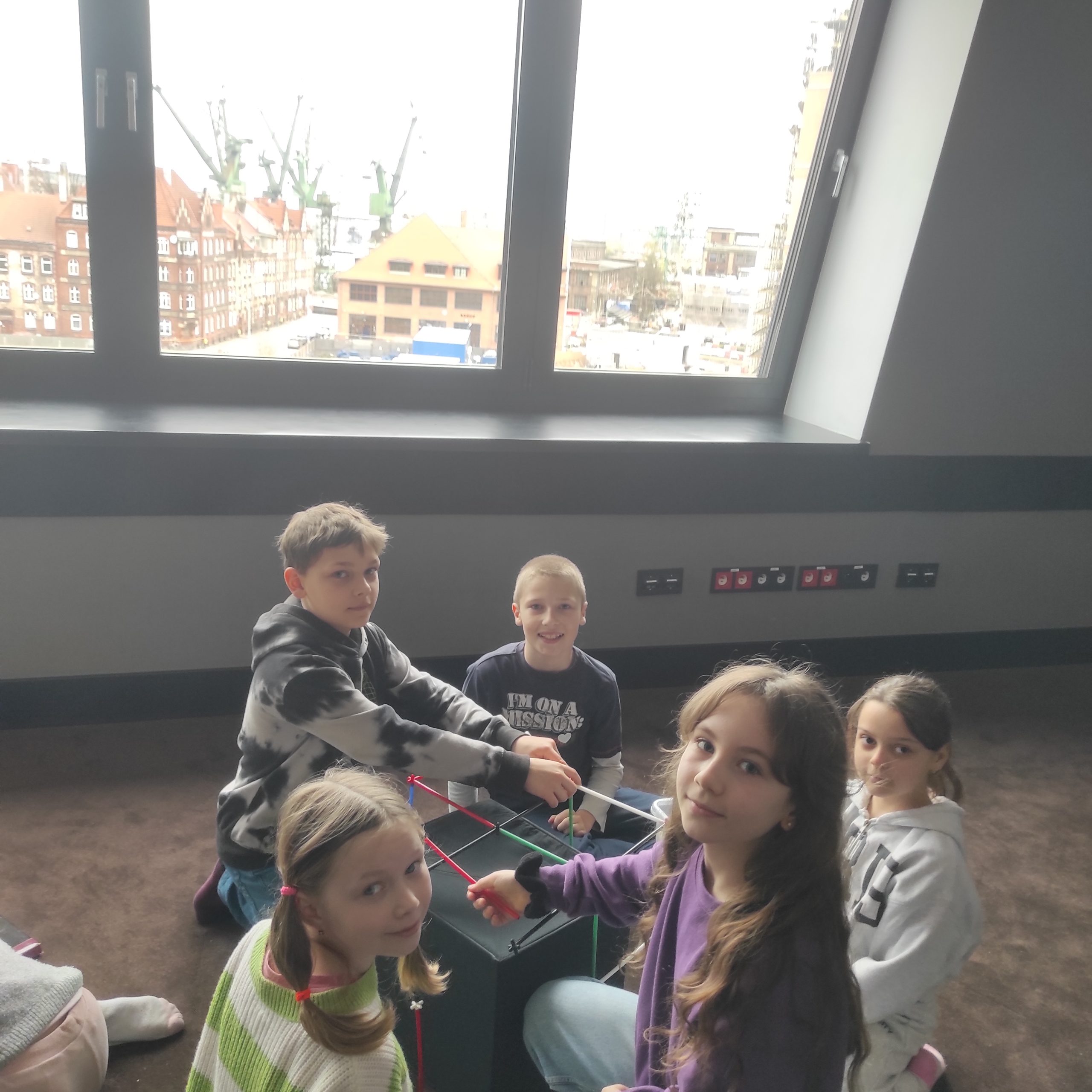 5 uczniów siedzi na podłodze wokół budowli z kolorowych rurek. W tle okno z widokiem na budynki i dźwigi.