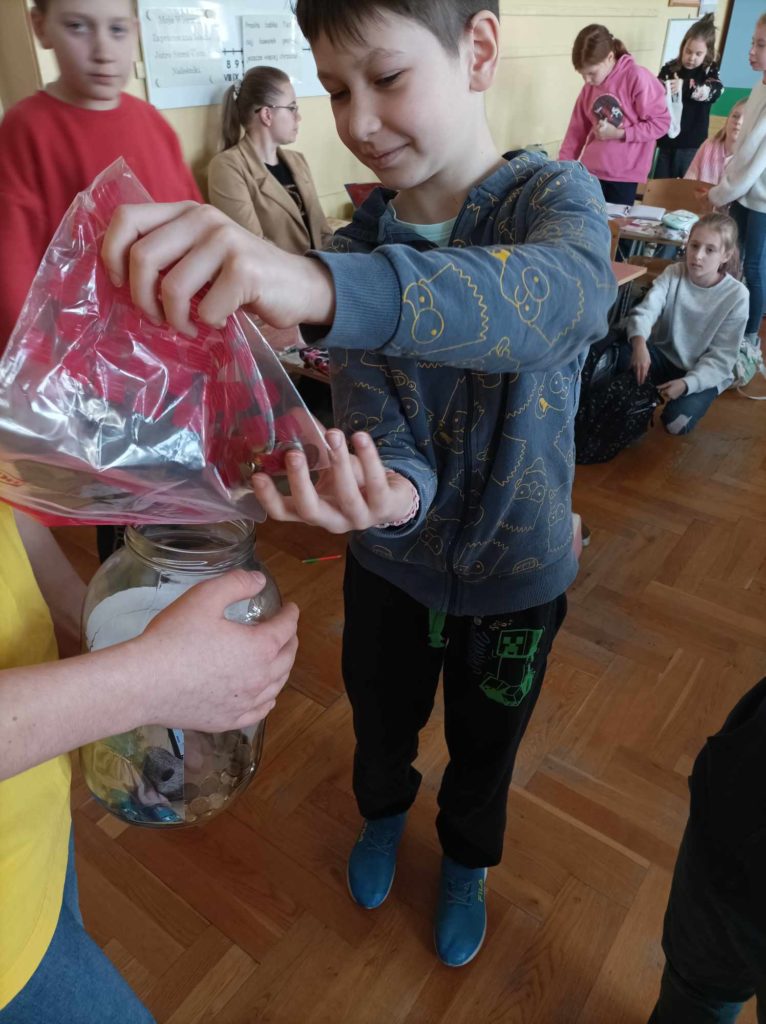 Chłopiec trzyma w ręku plastikowy woreczek, z którego przesypuje do dużego słoika grosze. Słoik trzyma drugi chłopiec. W tle są dzieci, które znajdują się w sali lekcyjnej.