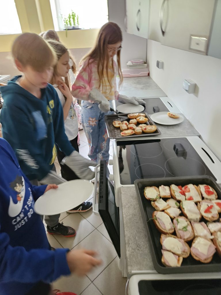 Na zdjęciu widzimy grupkę dzieci układajacy przygotowane zapiekanki do włożenia do piekarnika. Na blatach leżą blaszki, na których widzimy rozłożone kanapki