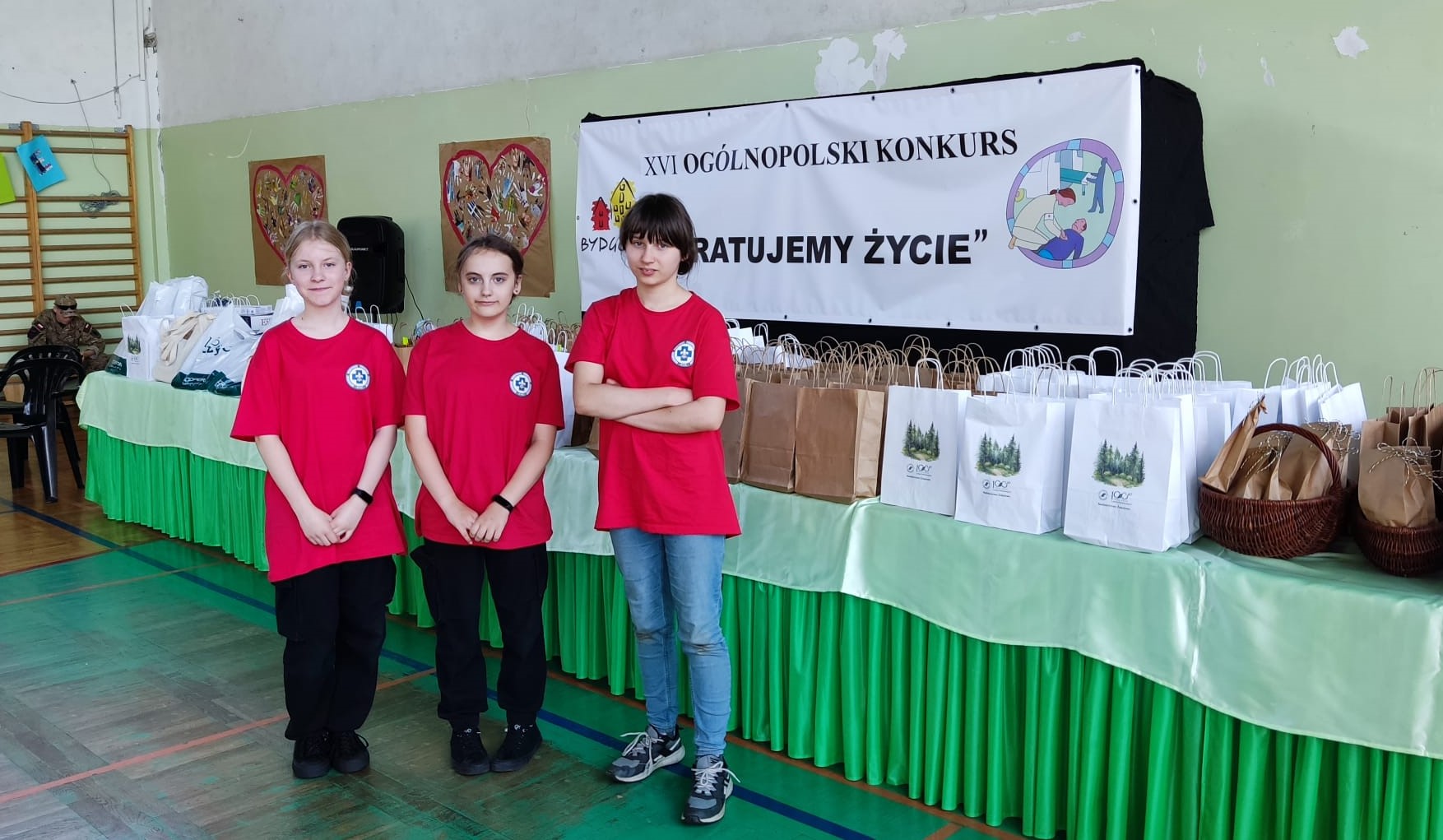 W sali gimnastycznej na tle plakatu XVI Ogólnopolskiego Konkursu „Ratujemy życie” i nagród ustawionych na zielonym stole stoją trzy uczennice SP 47 ubrane w czerwone koszulki. W tle na krześle siedzi uczeń szkoły mundurowej.