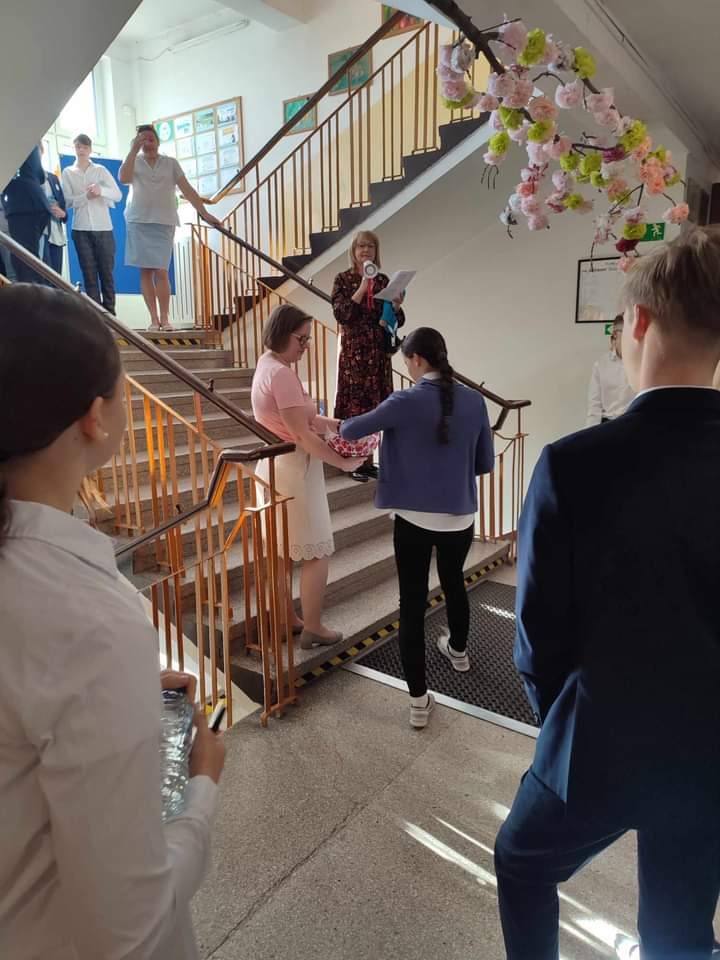Na szkolnym korytarzu na schodach stoją dwie kobiety, przed nimi obrócona tyłem uczennica sięga ręką do koszyka ze słodyczami. Na pierwszym planie dwoje uczniów widocznych z tyłu.