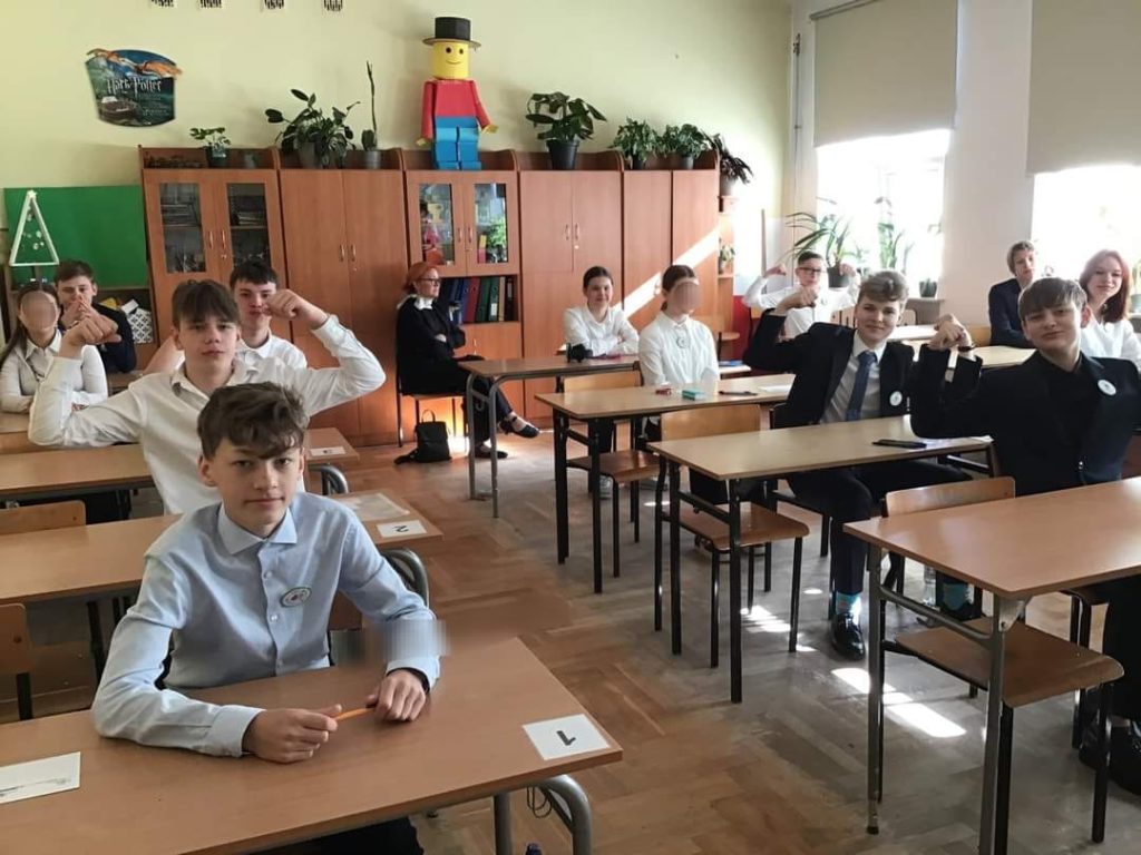 W sali lekcyjnej przy stołach siedzi 12 uczniów. Niektórzy z nich mają podniesione ręce w geście mocy (siły)
