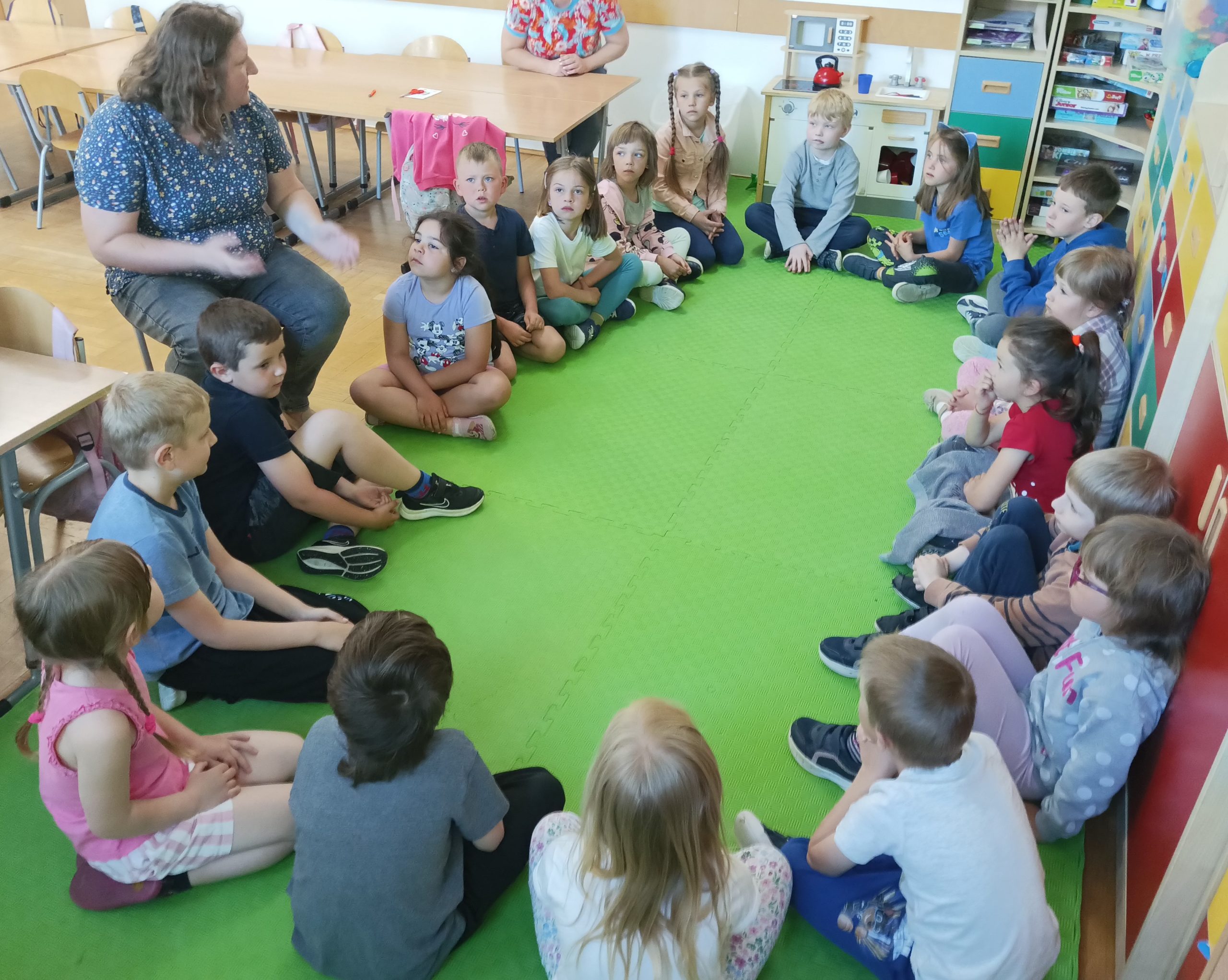 Na podłodze w klasie w kręgu siedzi grupa przedszkolaków i patrzą na kobietę, która siedzi na krześle. W tle wyposażenie szkolnej klasy.