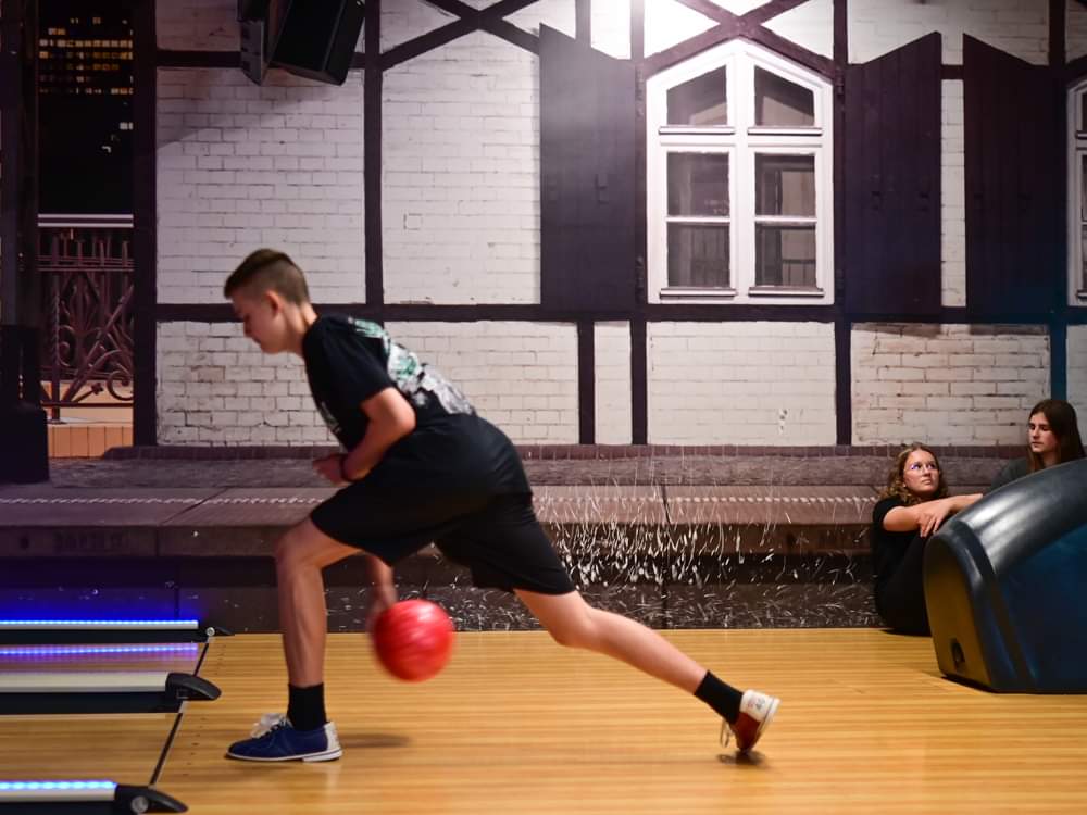 Uczeń z klasy 7a wykonuje rzut kulą na torze bowlingowym. W tle siedzą dwie dziewczynki.