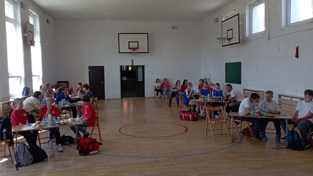 W sali gimnastycznej przy sześciu stolikach siedzą trzyosobowe drużyny wraz z opiekunami, obok stolików leżą apteczki. W tle dwie siedzące na krzesłach obok drzwi nauczycielki.