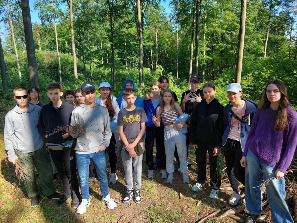 16 nastolatków stoi w lesie, niektórzy z nich trzymają w rękach patyki.