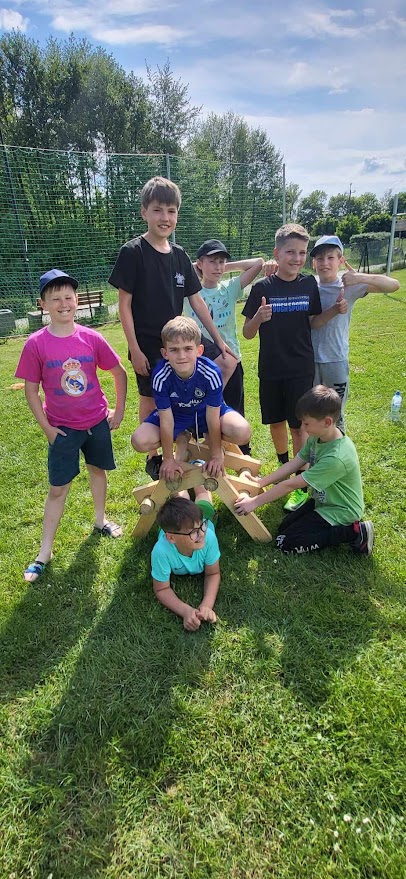 8 chłopców na boisku stoi lub kuca przy budowli z drewnianych elementów, jeden z chłopców leży na trawie pod budowlą.