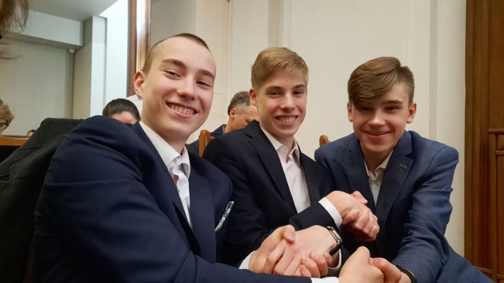 Trzech odświętnie ubranych ósmoklasistów uśmiecha się i trzyma się za ręce w geście gratulacyjnym. Chłopcy siedzą w gmachu Politechniki Gdańskiej.