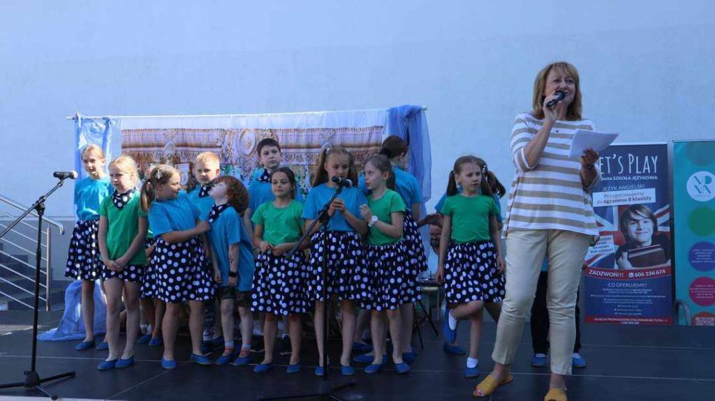 Na scenie kilkanaścioro śpiewających dzieci , z prawej strony z mikrofonem w dłoni Pani Dyrektor