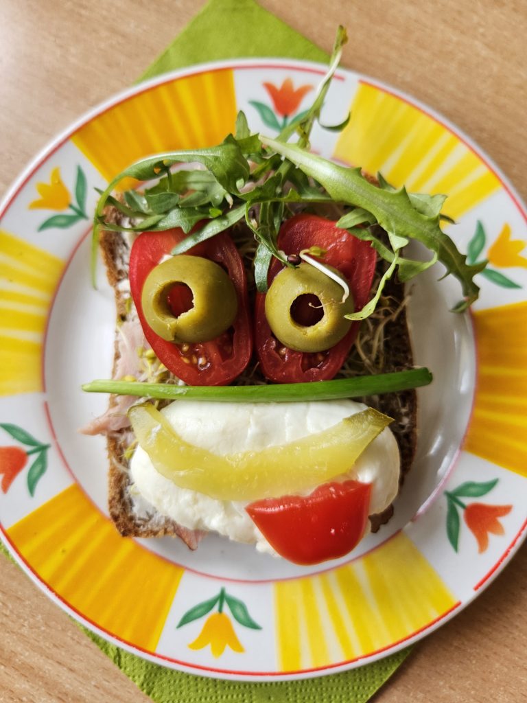 Na tacce „uśmiechnięta” kanapka udekorowana oliwkami, rukolą, pomidorkiem