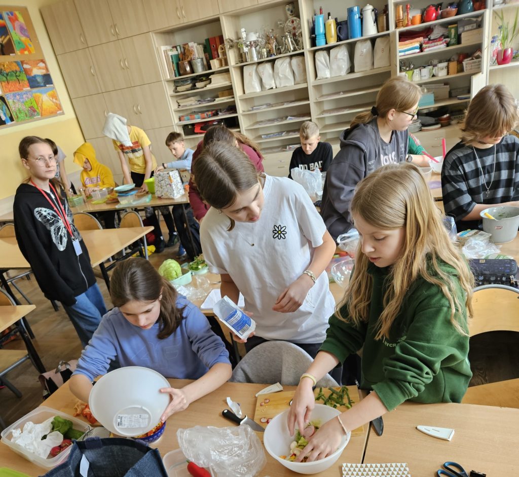 Na pierwszym planie stoły z porozkładanymi produktami. Uczniowie obierają i kroją warzywa