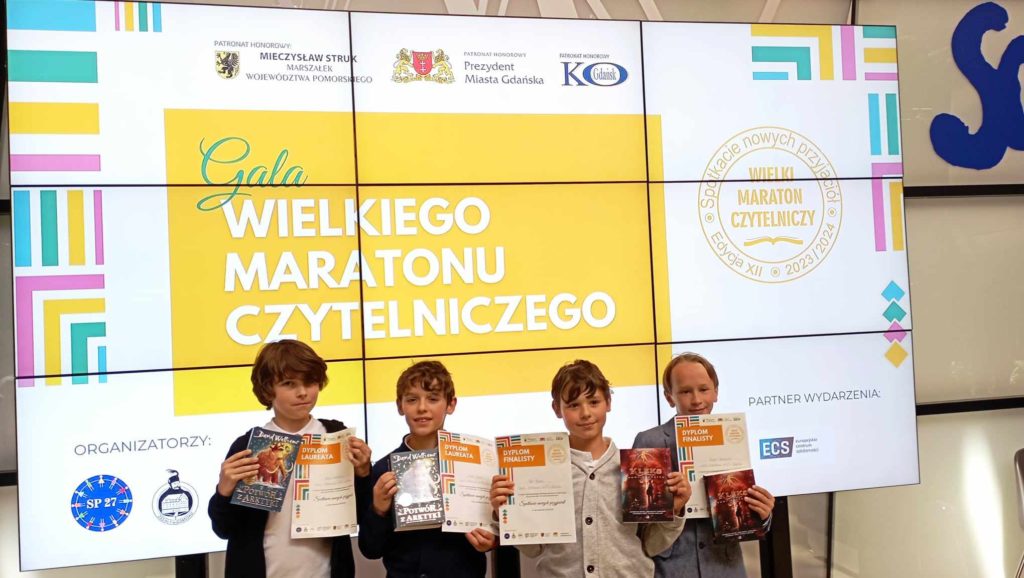Na tle telebimu z napisem Gala Wielkiego Maratonu Czytelniczego stoi czterech uczniów klas młodszych. W rękach trzymają dyplomy i nagrody książkowe.