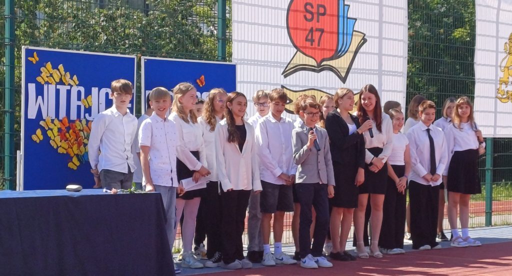 Uśmiechnięci uczniowie klasy 6b stoją w dwuszeregu na boisku szkolnym. Jeden chłopiec i jedna dziewczynka trzymają w rękach mikrofon.