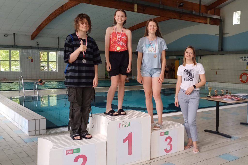 Trzy dziewczynki stoją na podium na pływalni. Na szyjach mają zawieszone medale. Po prawej stronie stoi pani dyrektor.