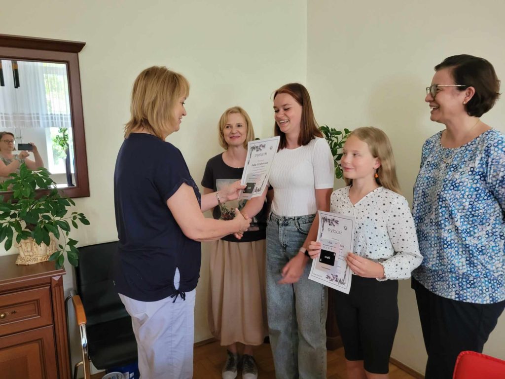 Uczennica odbiera dyplom i nagrodę z rąk dyrektora szkoły, obok stoi młodsza uczennica oraz dwie nauczycielki.