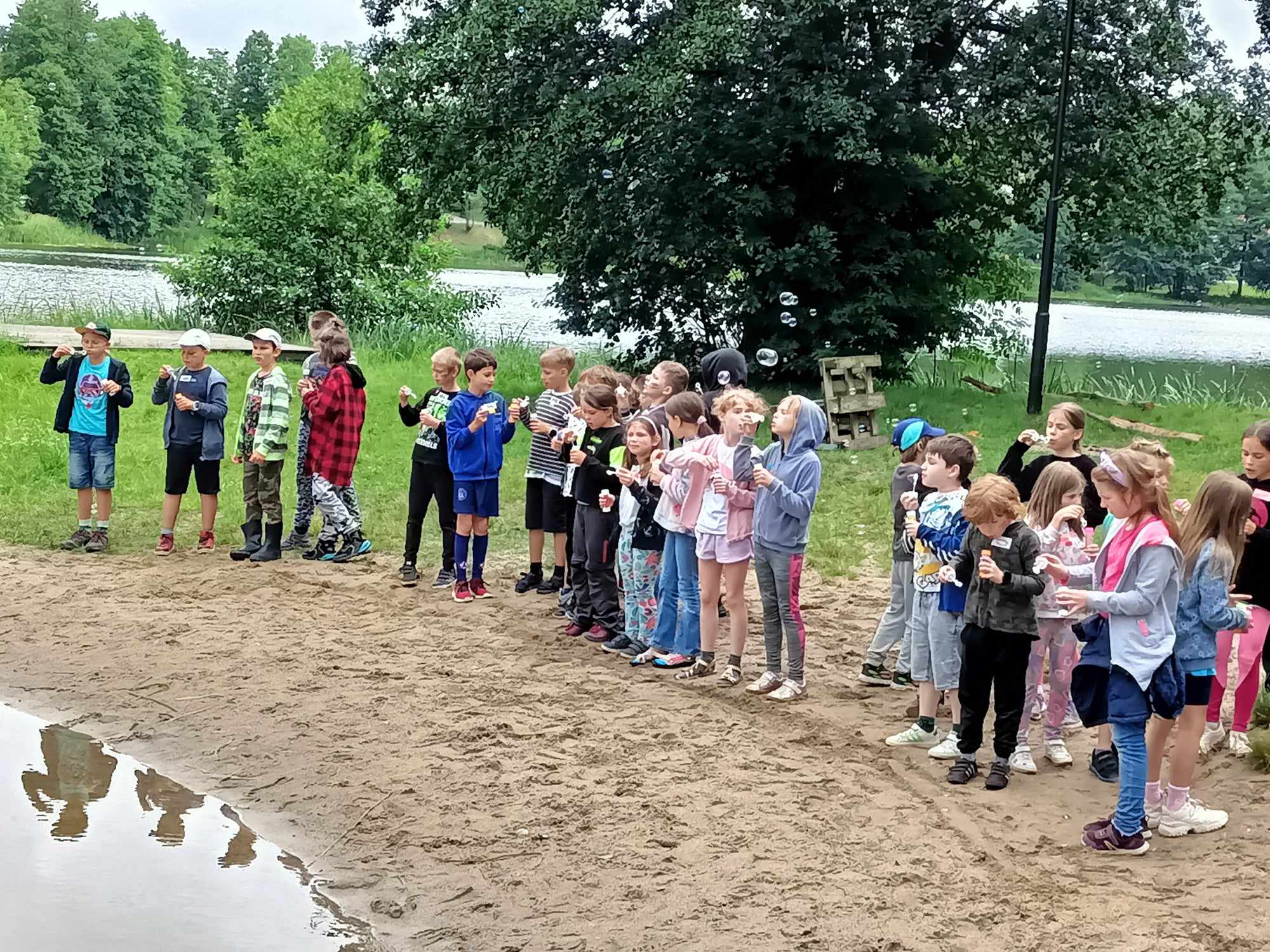 Grupa 26 uczniów stoi na piaszczystej plaży nad jeziorem. Dzieci dmuchają bańki mydlane, które unoszą się nad nimi. W tle rosną drzewa i krzewy.