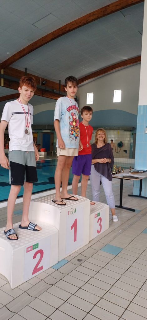 Trzech uśmiechniętych chłopców stoi na podium na basenie. Na szyjach mają zawieszone medale. Obok podium, po prawej stronie stoi pani dyrektor.