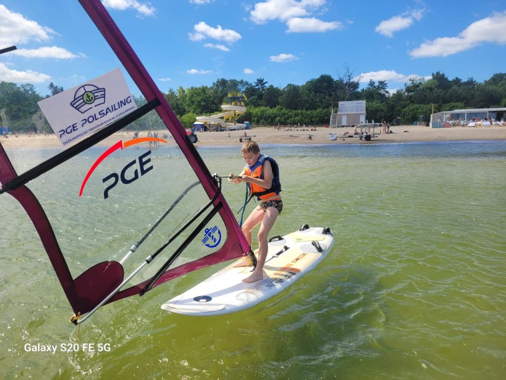 chłopiec stoi na desce windsurfigowej na wodzie trzyma w rękach żagiel