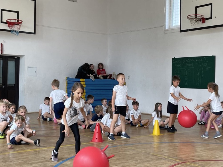Uczniowie klas I na sali gimnastycznej rywalizują w zawodach sportowych - wyścigi rzędów, dzieci skaczą na piłce