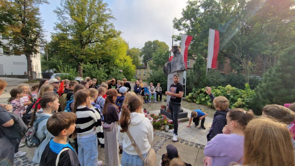 Grupa kilkudziesięciu uczniów zgromadzona wokół pomnika Danuty Siedzikówny. Słuchają opowiadania nauczyciela, poznając historię dzielnej Polki.