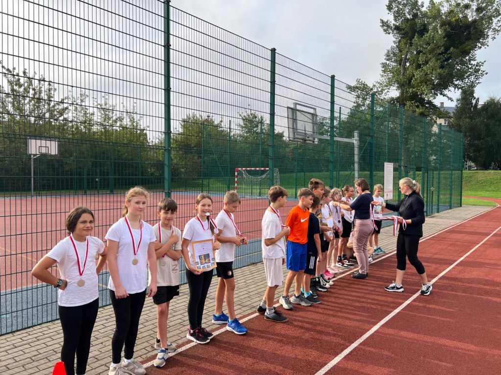 Uczniowie klas piątych stoją w szeregu na boisku szkolnym. Pani dyrektor dekoruje ich medala-mi. Obok stoi pani nauczycielka i trzyma w rękach tacę z medalami.