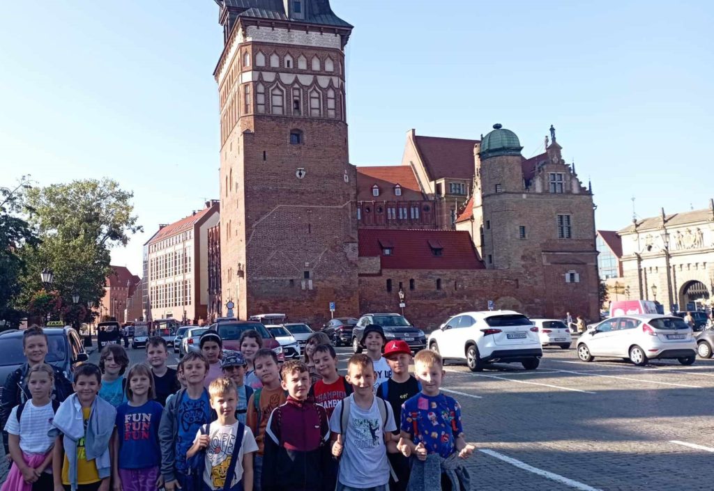 Na parkingu stoją w trzech rzędach uczniowie klasy 3c wraz z wychowawczynią. W tle gdański zabytek Katownia i zaparkowane na parkingu samochody.