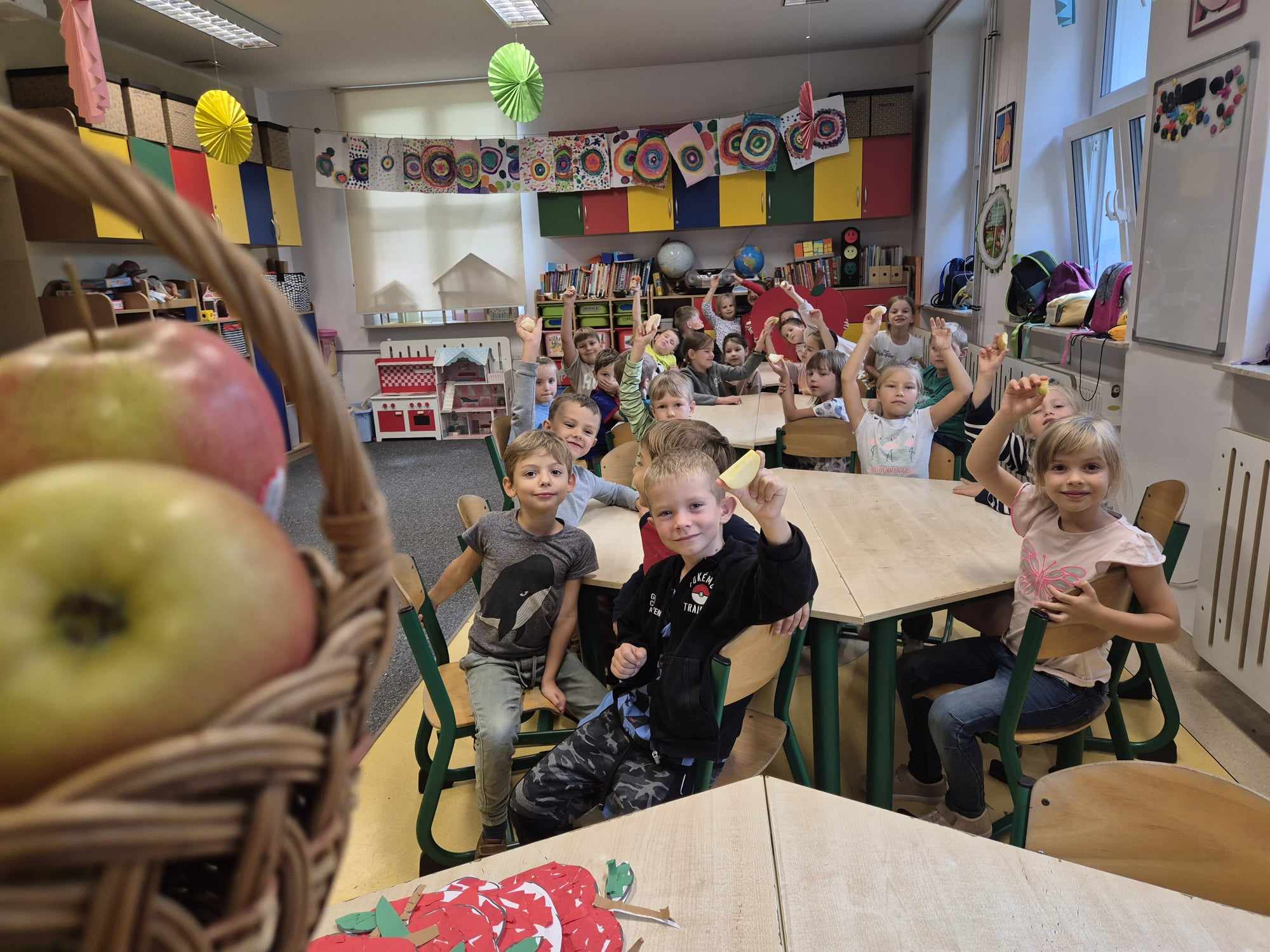 Na pierwszym planie kosz wiklinowy z jabłkami. W tle dzieci siedzą przy stoliku i jedzą jabłka.