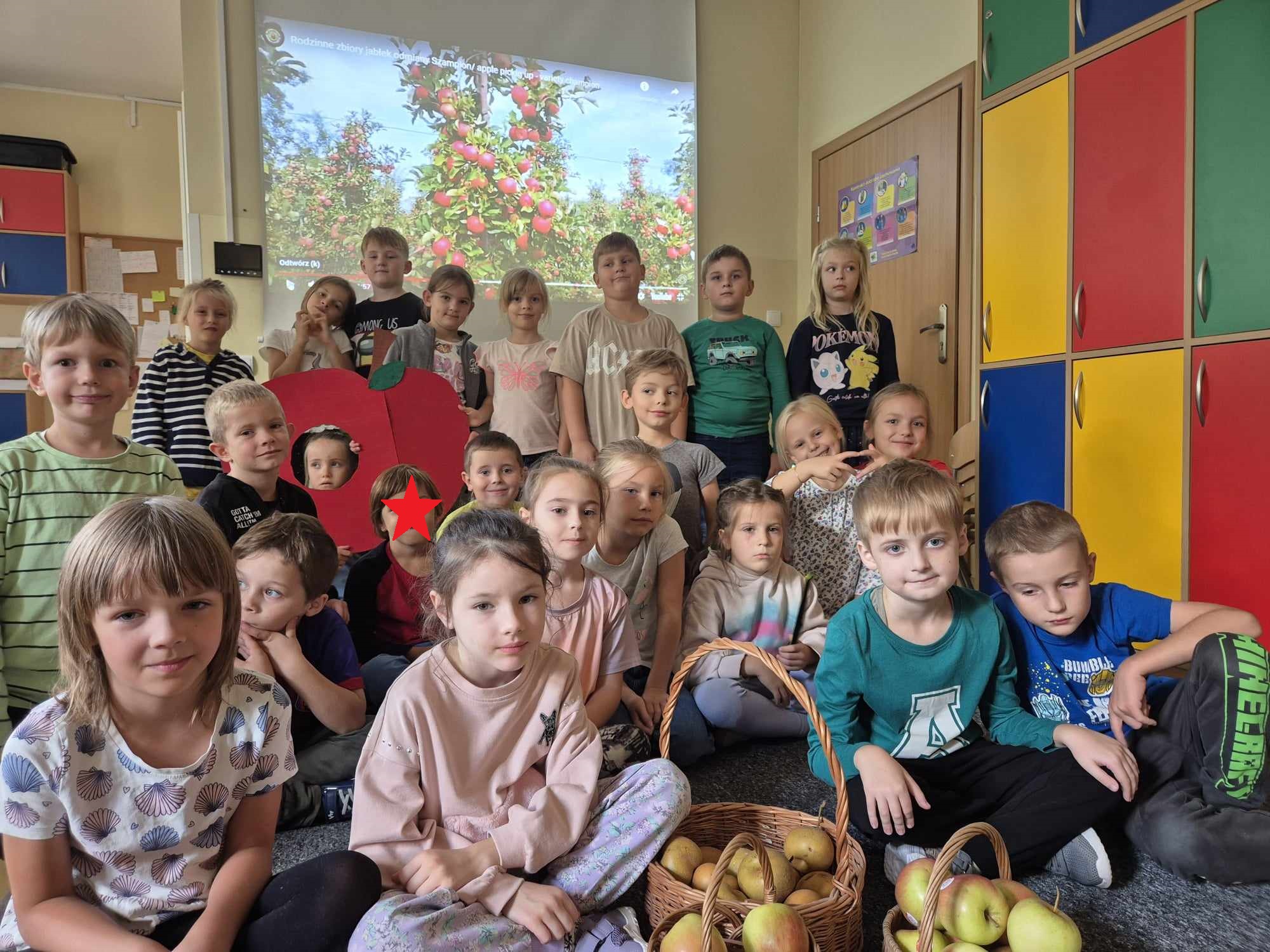 Grupa dzieci znajduje się w sali świetlicowej. Za nimi jest ekran, na którym wyświetlono są drzewa jabłoni. Przed dziećmi, na podłodze stoją kosze wiklinowe z jabłkami.