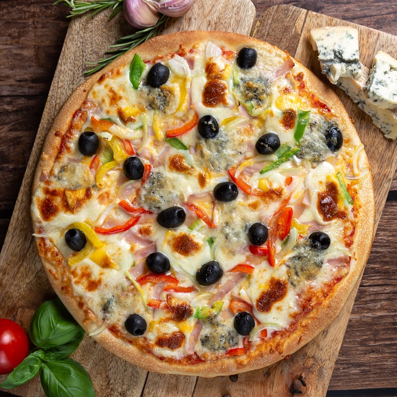 Upieczona okrągła pizza, z serem szynką, czerwoną papryką i oliwkami czarnymi leży na drewnianej desce . U góry po lewej lezą na desce 2 ząbki czosnku, po prawej kawałek sera pleśniowego. Na dole po lewej leży mały pomidor z bazylią.