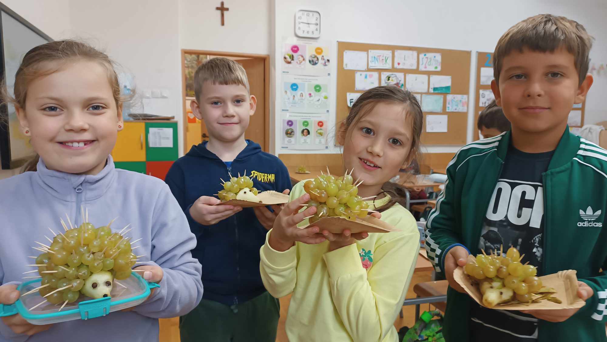  Klasa szkolna, dwie dziewczynki i dwóch chłopców stoi trzymając w ręku talerzyki, na których jest jeżyk, wykonany z gruszki i winogron nakłutych na wykałaczki. W tle od lewej strony kolorowe szkolne ławki, drzwi, zegar na ścianie i tablice korkowe z rysunkami.