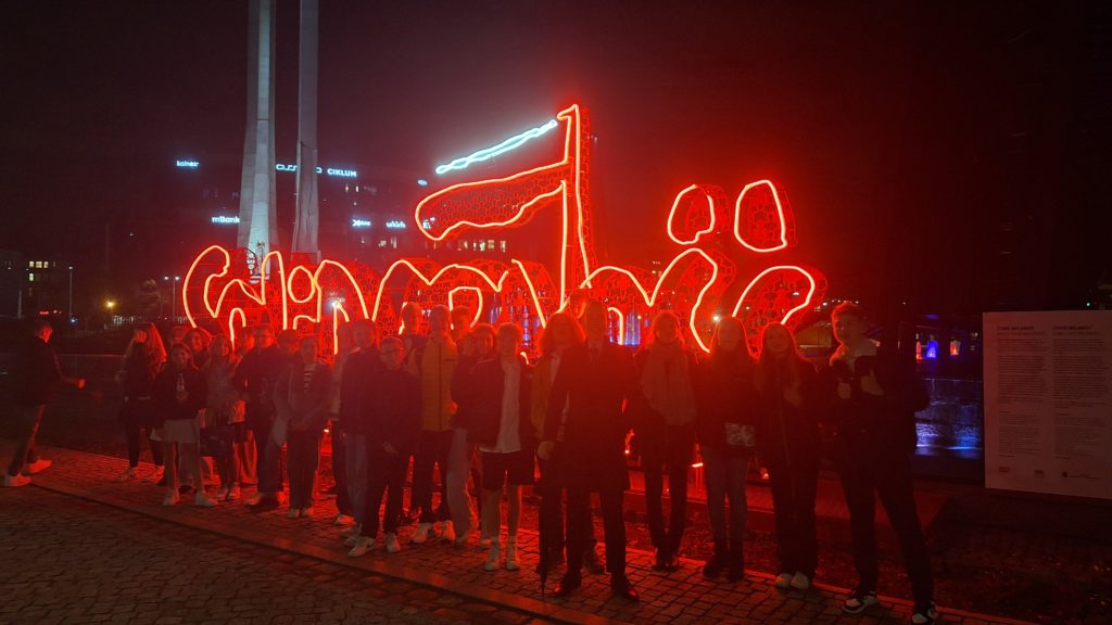 kilkunastu młodych osób stoi na tle neonowego napisu solidarność. Jest ciemno i postacie są niewyraźne