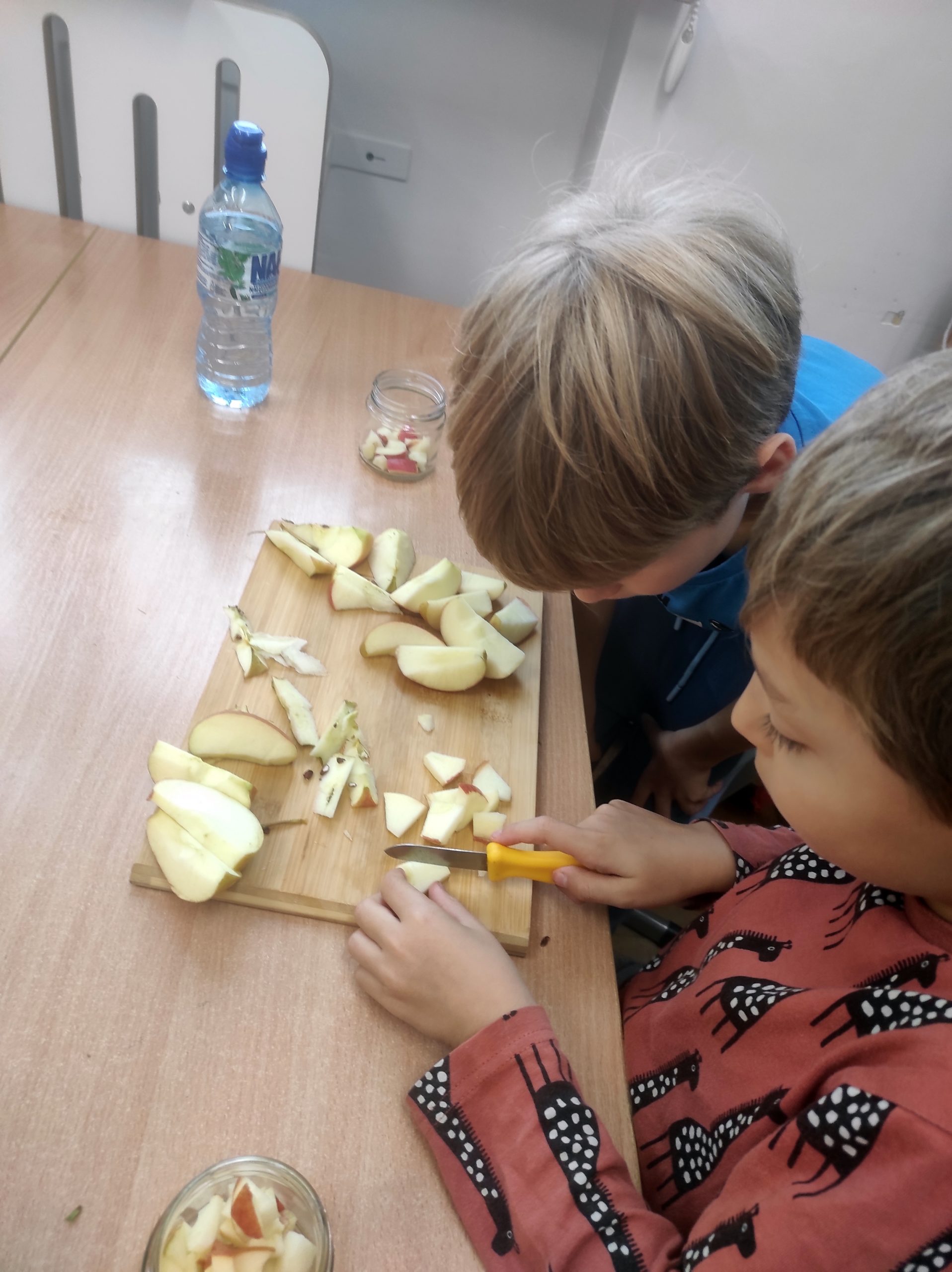 Po prawej stronie stolika siedzi 2 chłopców, którzy są pochyleni nad deską do krojenia, na której są cząstki jabłka. Po obu stronach deski stoją słoiki wypełnione cząstkami jabłka. Obaj chłopcy są pochyleni nad deską, pierwszy chłopiec kroi jabłko nożykiem, drugi przygląda się temu.