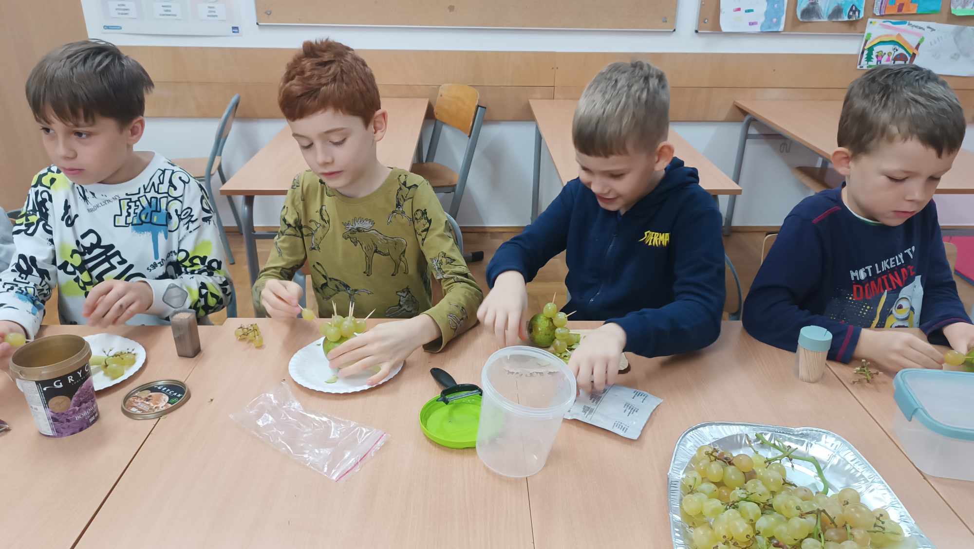 Klasa szkolna, na przodzie duży stół. Za stołem siedzi czworo chłopców wykonujących jeża z owoców, na białych talerzykach. Na stole na tacy winogrona oraz plastikowe okrągłe pudełka, woreczek foliowy, wykałaczki. Za uczniami w rzędzie stoją cztery ławki szkolne z krzesełkami. W tle od lewej drzwi i dwie tablice z obrazkami.