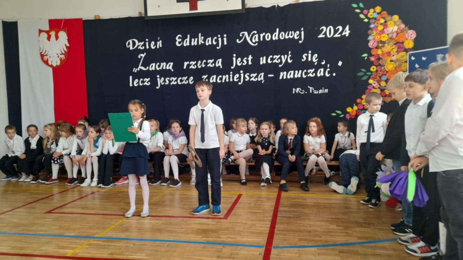 Aula szkolna, z tyłu na granatowym tle napis: Dzień Edukacji Narodowej 2024, Zacną rzeczą jest uczyć się, lecz jeszcze zacniejszą- nauczać.'' Po lewej stronie godło Polski na biało-czerwonej fladze. Z przodu stoi dziewczynka i chłopiec, za nimi siedzą dzieci w dwóch rzędach na ławeczkach. Po prawej stronie stoją dzieci ubrane na galowo. Wszyscy ubrani na galowo.