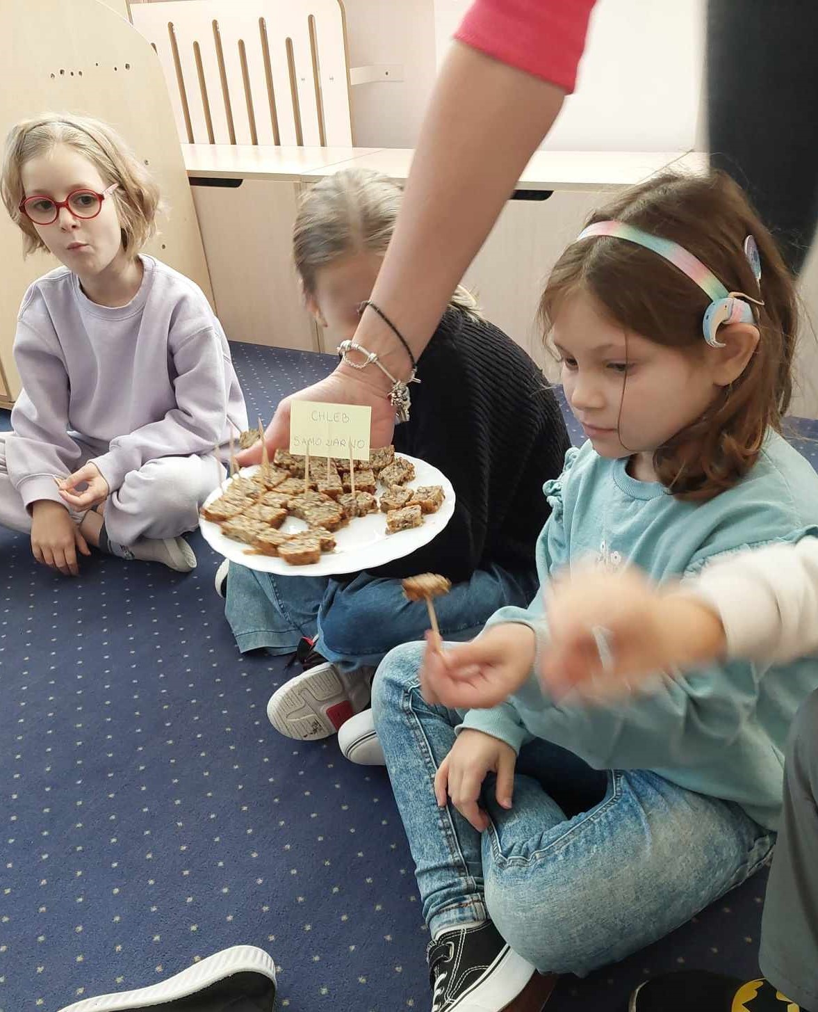 3 dziewczynki siedzą na dywanie. Po między nimi ręka nauczycielki trzyma talerz z pokrojonym chlebem. 