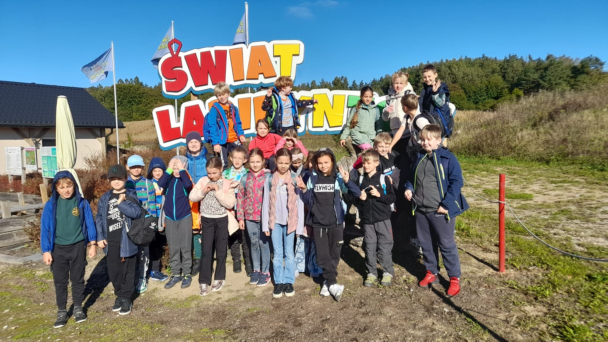 Dwudziestoosobowa grupa dzieci stoi przed wejściem na plac labiryntów. Nad nimi jest czerwono-pomaranczowo-żółty napis ŚWIAT LABIRYNTÓW.