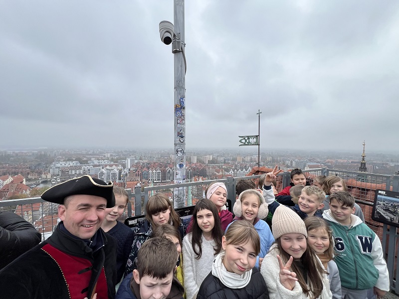 Uczniowie ustawieni w grupce jeden obok drugiego na punkcie widokowym Bazyliki Mariackiej a z lewej strony pirat Paweł. Za nimi rozpościera się panorama Gdańska