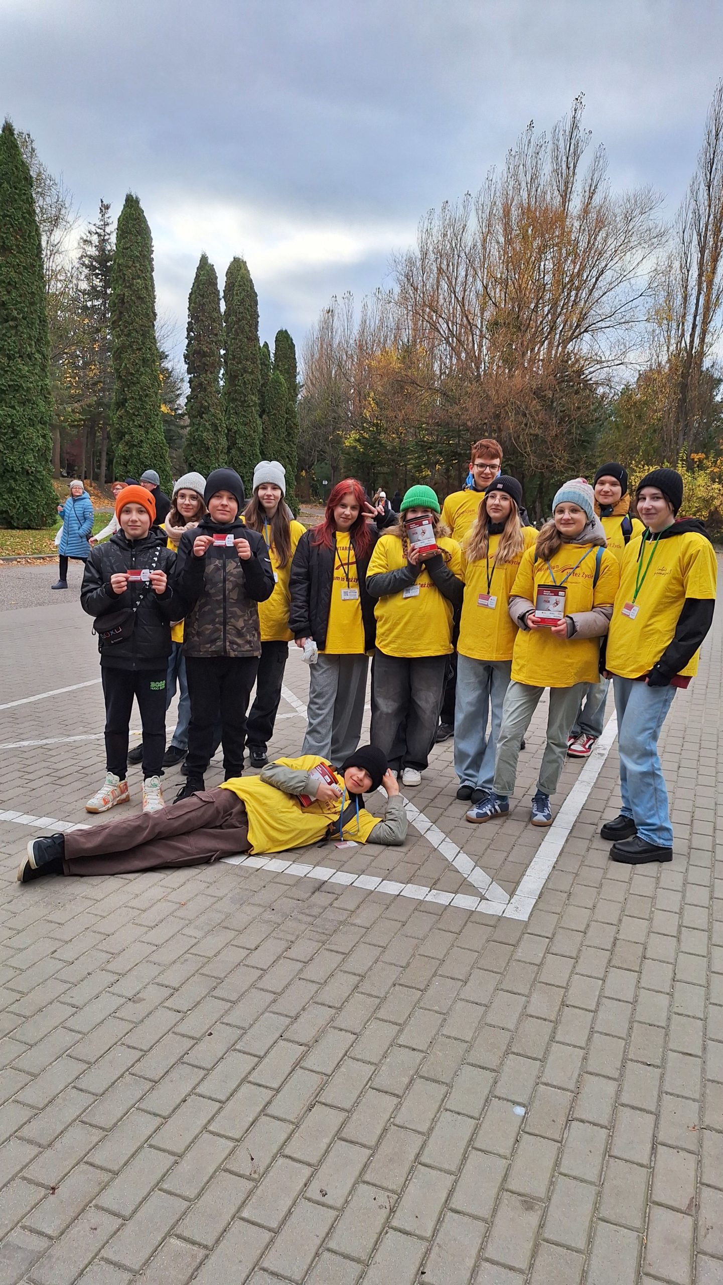  cmentarz - Grupa młodych osób ubrana w koszulki wolontariuszy stoi na tle drzew. Jeden chłopiec leży przed grupą na chodniku opierając głowę o rękę. Niektóre osoby trzymają w ręku skarbonki.