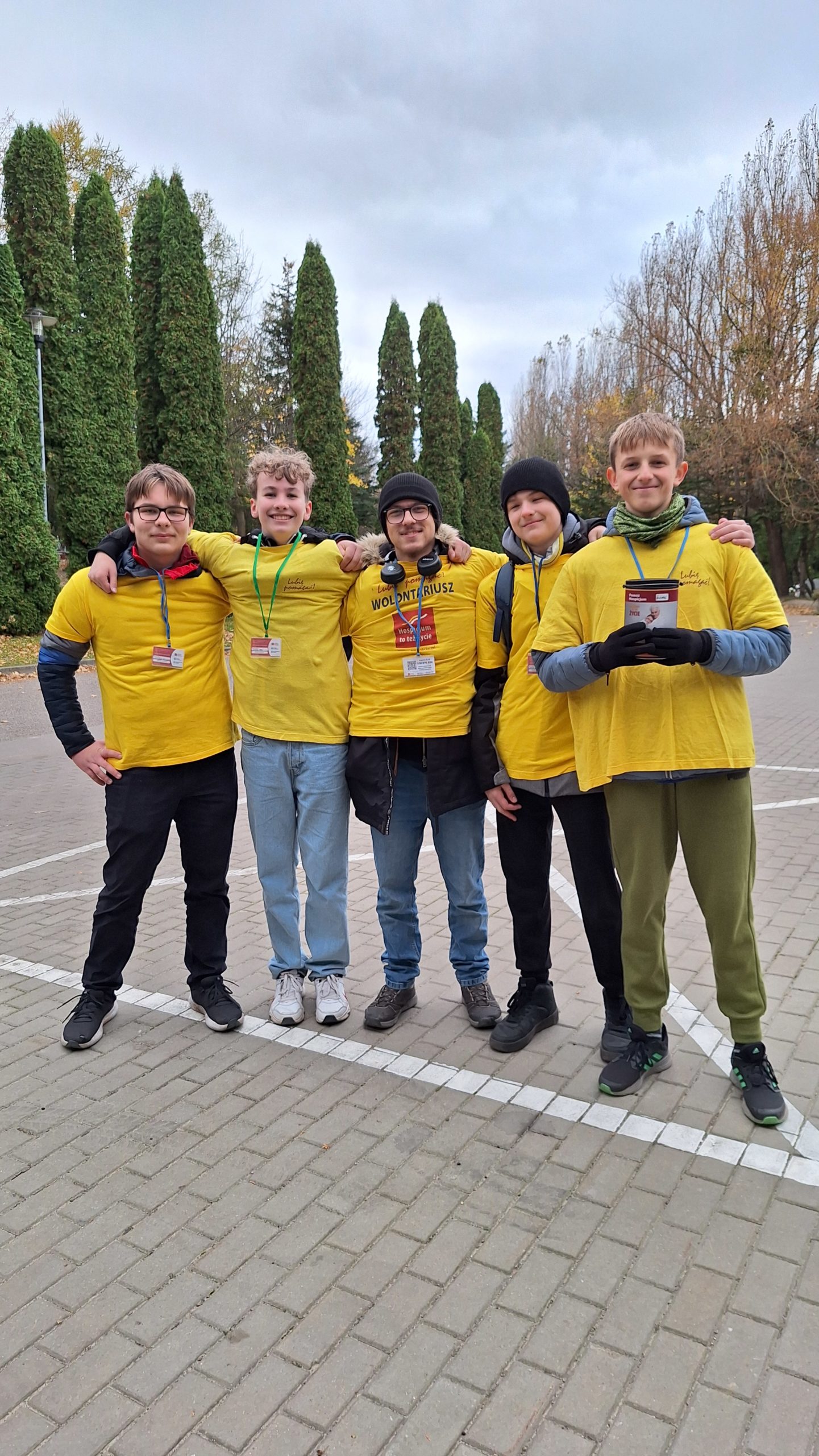 cmentarz - 5 chłopców stoją w rzędzie na tle drzew i trzymają się za barki. Ubrani są w koszulki wolontariuszy, a na szyi mają zawieszone identyfikatory. Jeden z nich w rękach trzyma puszkę na pieniądze. Chłopcy są uśmiechnięci 