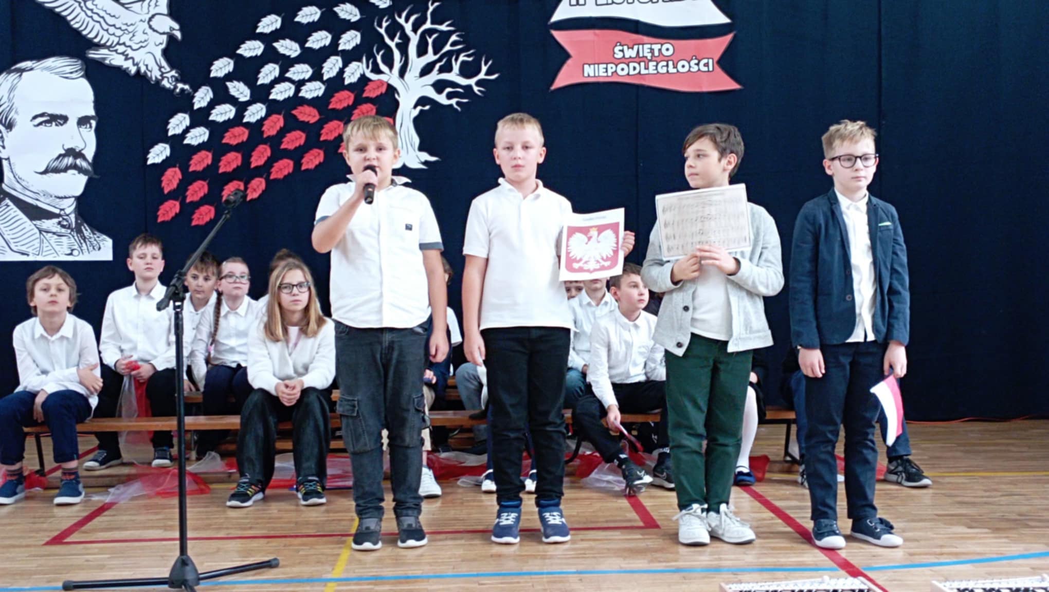 W sali gimnastycznej stoi czterech uczniów z klasy 3c, jeden trzyma mikrofon, drugi godło, trzeci nuty humnu, a czwarty flagę. Przed nimi mikrofon na statywie. W tle dzieci siedzące na ławeczkach, dekoracja z orłem, wizerunkiem Piłsudskiego, drzewem z białymi i czerwonymi liśćmi oraz napisem 11 listopada Święto Niepodległości.