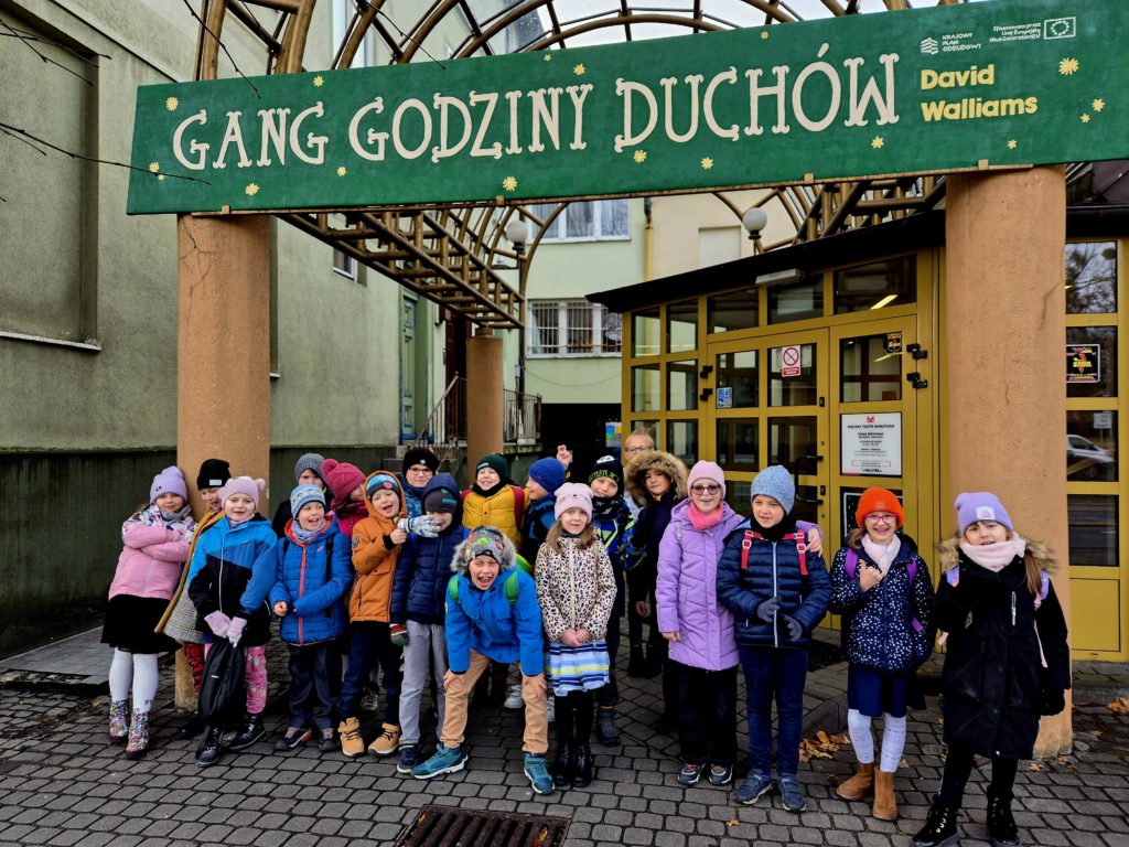 Przed budynkiem Teatru Miniatura stoi dwudziestka dzieci. Jeden chłopiec w środku grupy kuca i ręce trzyma na kolanach. Dzieci są ubrane na zimowo. Nad dziećmi jest napis ,,Gang Godziny Duchów".