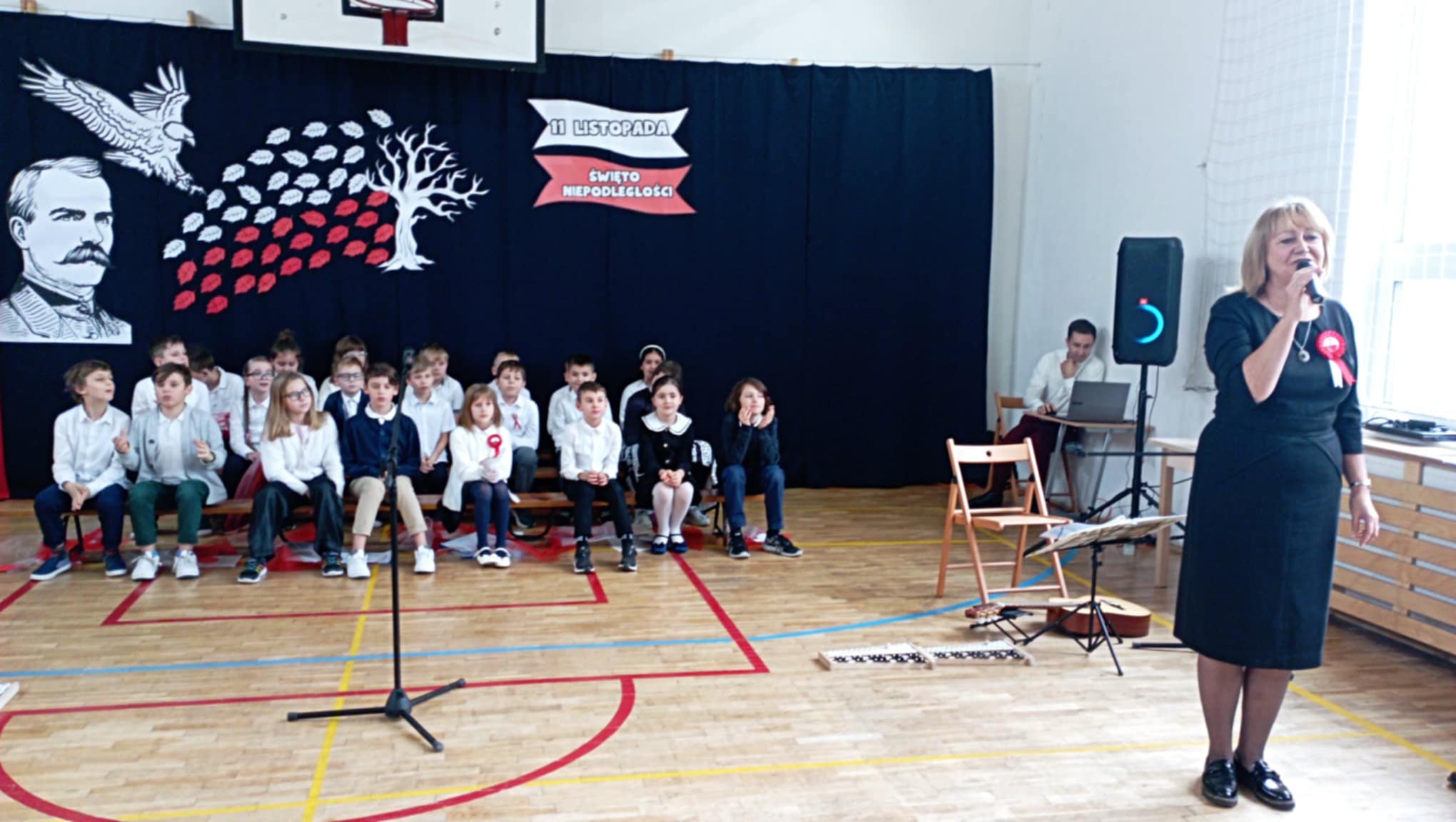 W sali gimnastycznej na pierwszym planie Dyrektor Szkoły i mikrofonem. Obok na podłodze leżą dzwonki i gitara. Za Dyrektorem uczniowie siedzący na ławeczkach i nauczyciel przy laptopie. W tle dekoracja z orłem, wizerunkiem Piłsudskiego, drzewem z białymi i czerwonymi liśćmi oraz napisem 11 listopada Święto Niepodległości.